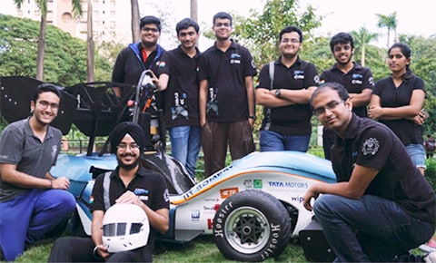 A group photo of twelve IIT Bombay students stands around a Formula-style racing car in a park. They wear matching black polo shirts and appear enthusiastic. The racing car in blue and white car features sponsor logos, and two students in the foreground are seated, one holding a white racing helmet, with lush greenery in the background.